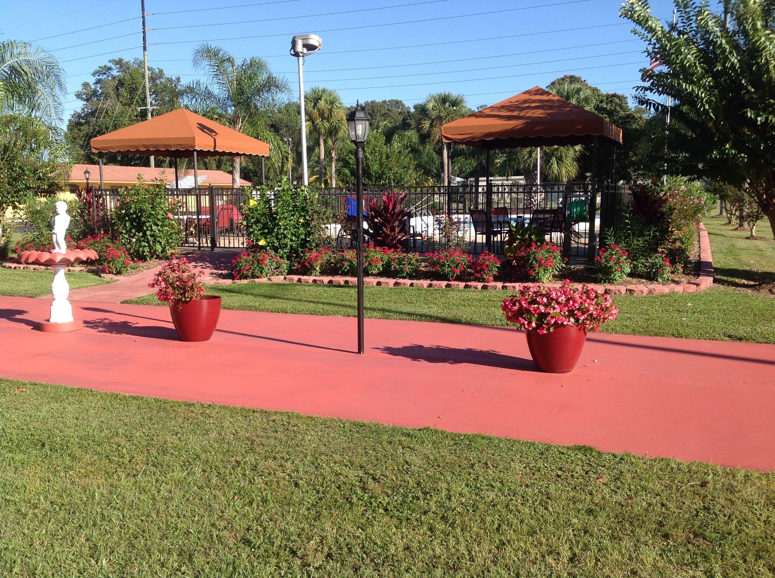 A garden with a red pathway, two large potted plants, a white statue, and a gazebo with a brown roof.