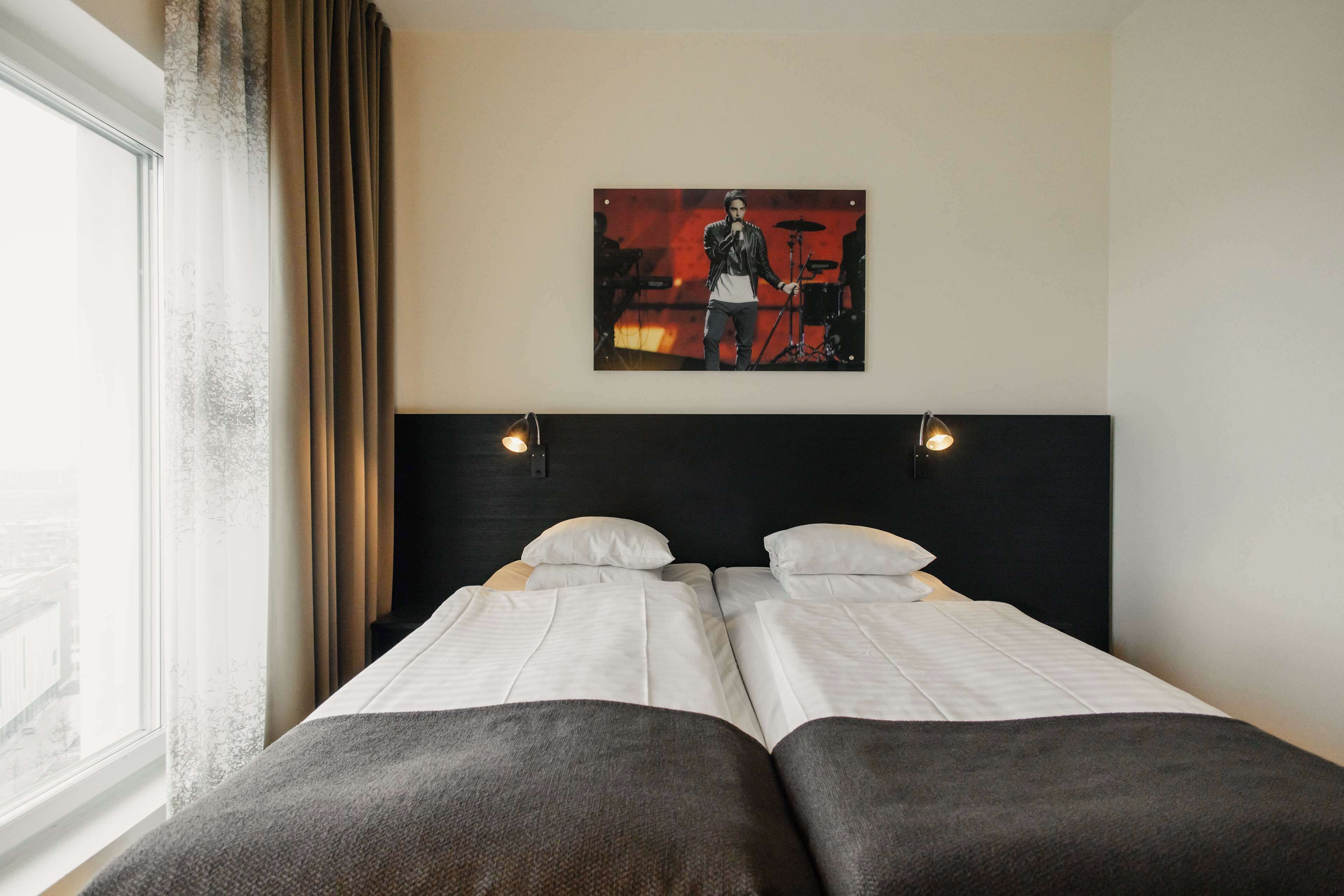 A hotel room with two single beds, a large window with curtains, a wall-mounted artwork, and a headboard with two wall-mounted lights.