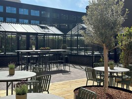 Un patio al aire libre con mesas y sillas, una estructura con paredes de vidrio y un árbol.