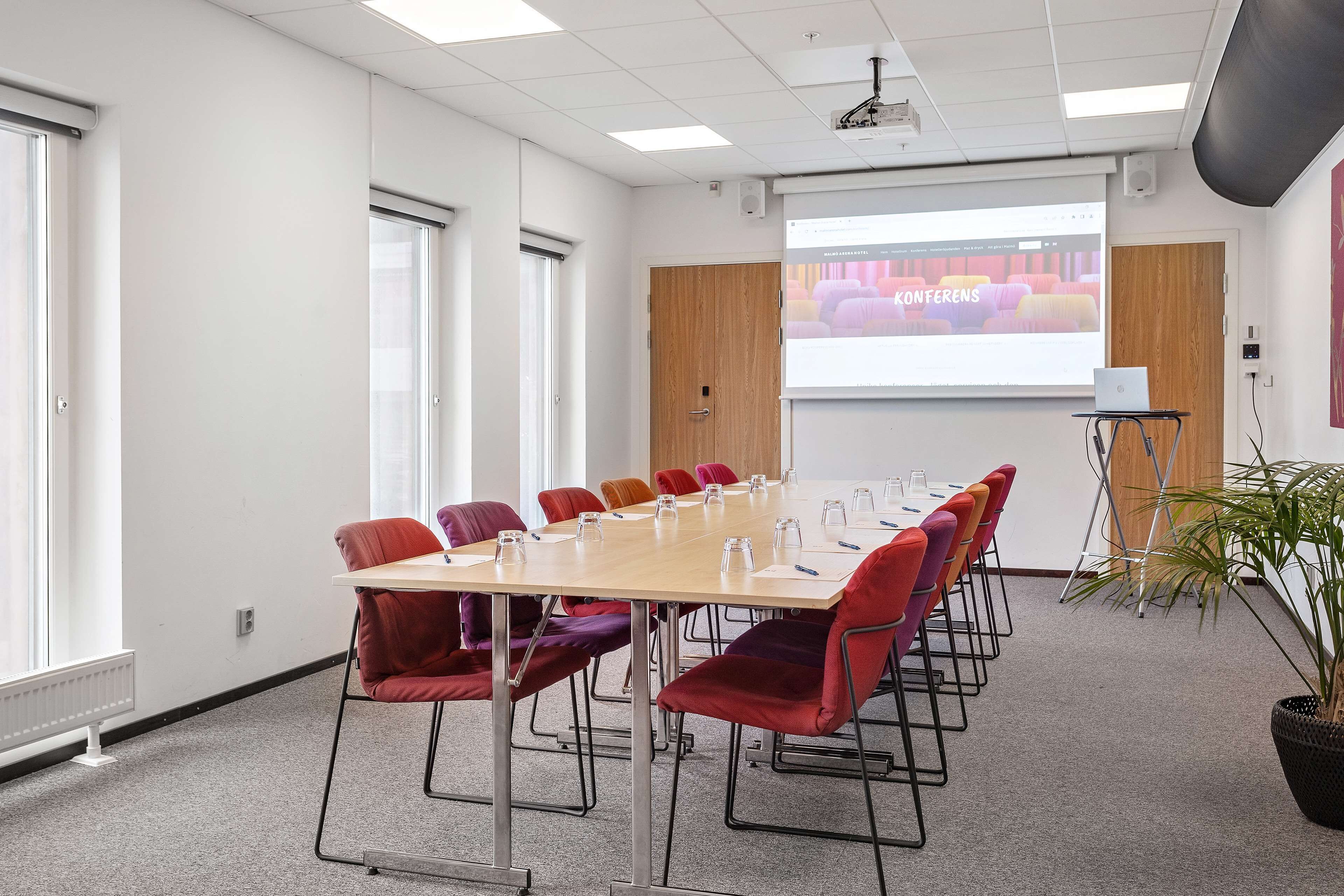 Ett konferensrum med ett långt bord, röda stolar, en projektorduk och en bärbar dator.