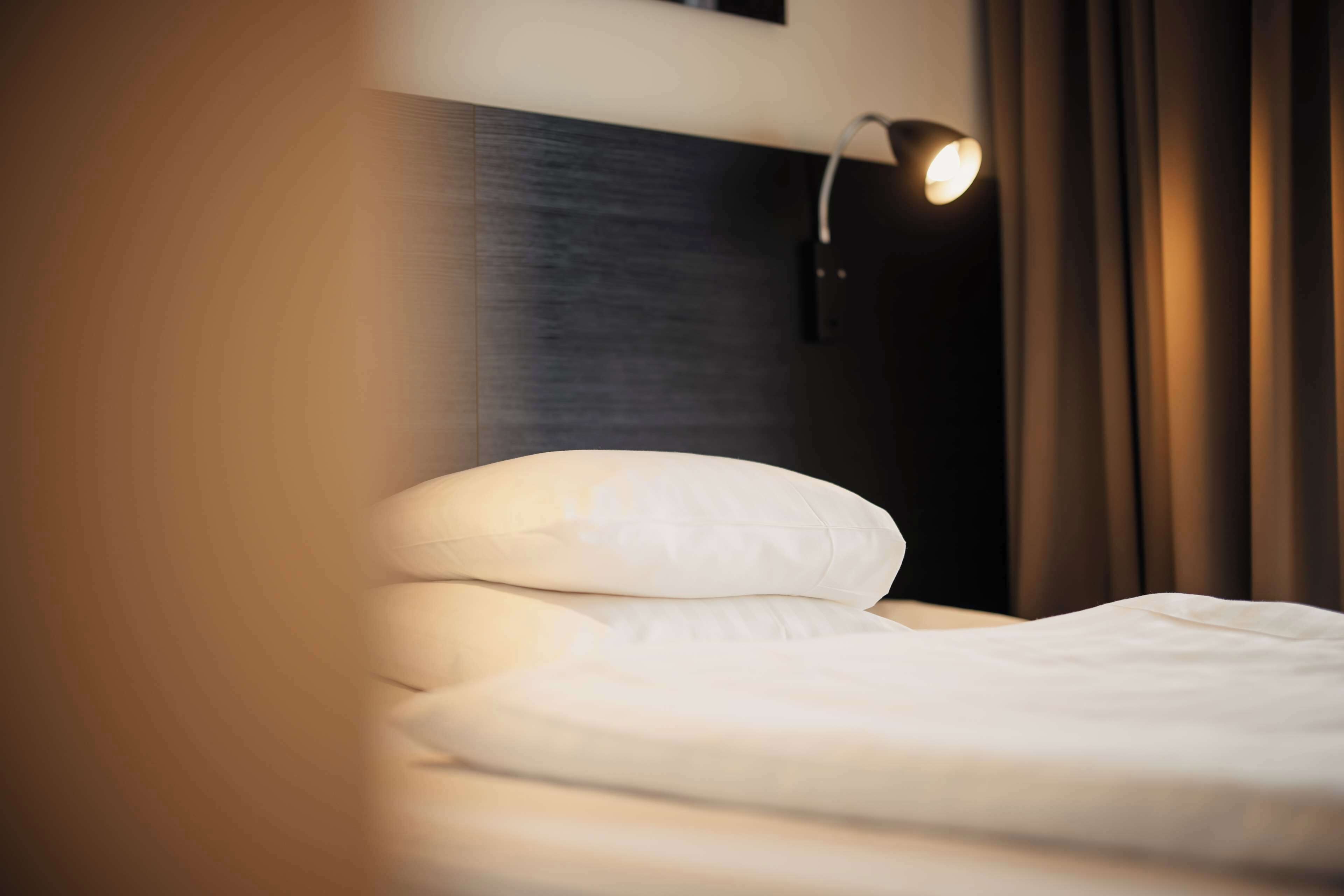 A neatly made bed with white bedding, a dark headboard, and a wall-mounted light fixture.