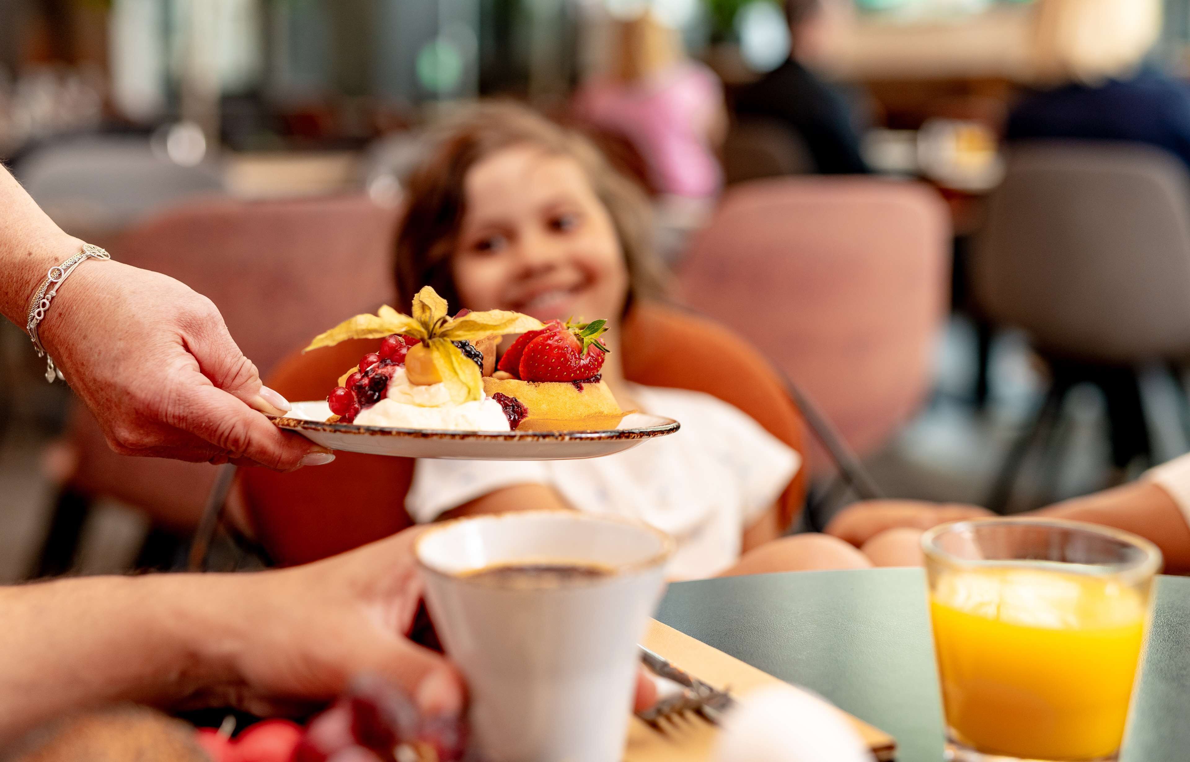 Eine Person serviert ein Dessert mit Erdbeeren und Sahne an ein Kind, das an einem Tisch mit Orangensaft sitzt.
