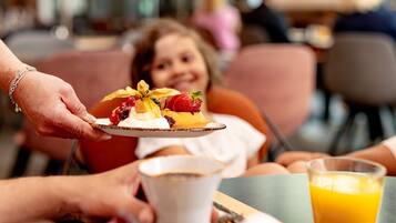 Eine Person serviert ein Dessert mit Erdbeeren und Sahne an ein Kind, das an einem Tisch mit Orangensaft sitzt.