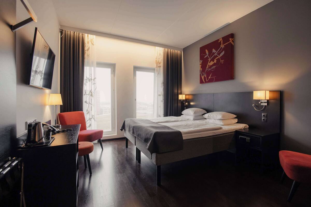 Une chambre d’hôtel moderne, équipée d’un lit, d’un bureau et de chaises. On y trouve des rideaux, un téléviseur et un tableau au mur.