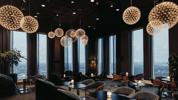 Un moderno vestíbulo de hotel con mesas redondas, asientos cómodos, ventanas amplias y lámparas decorativas colgantes.