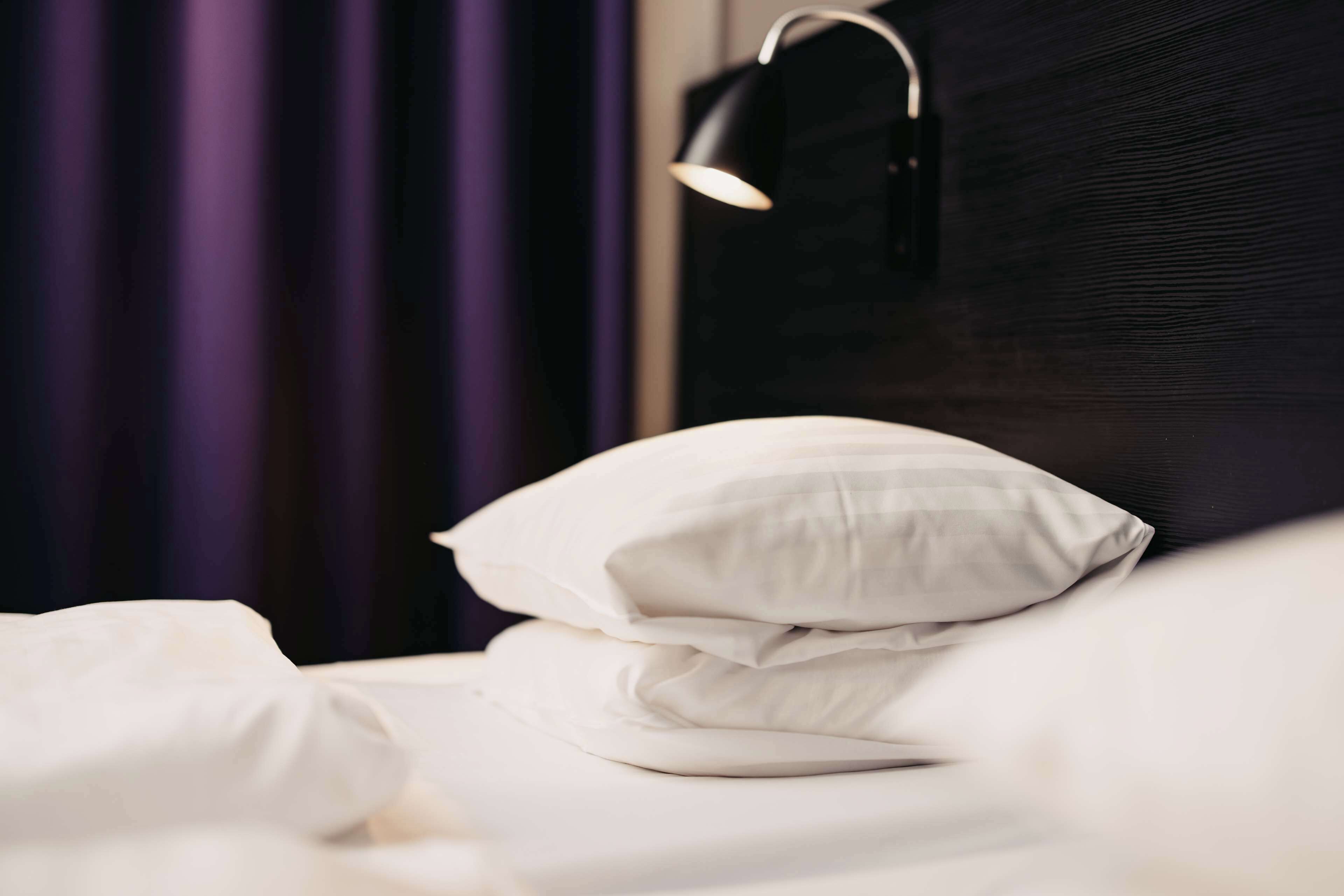 A neatly made bed with white bedding and a single white pillow, a black wall, and a purple curtain in the background.