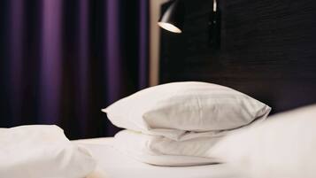 A neatly made bed with white bedding and a single white pillow, a black wall, and a purple curtain in the background.
