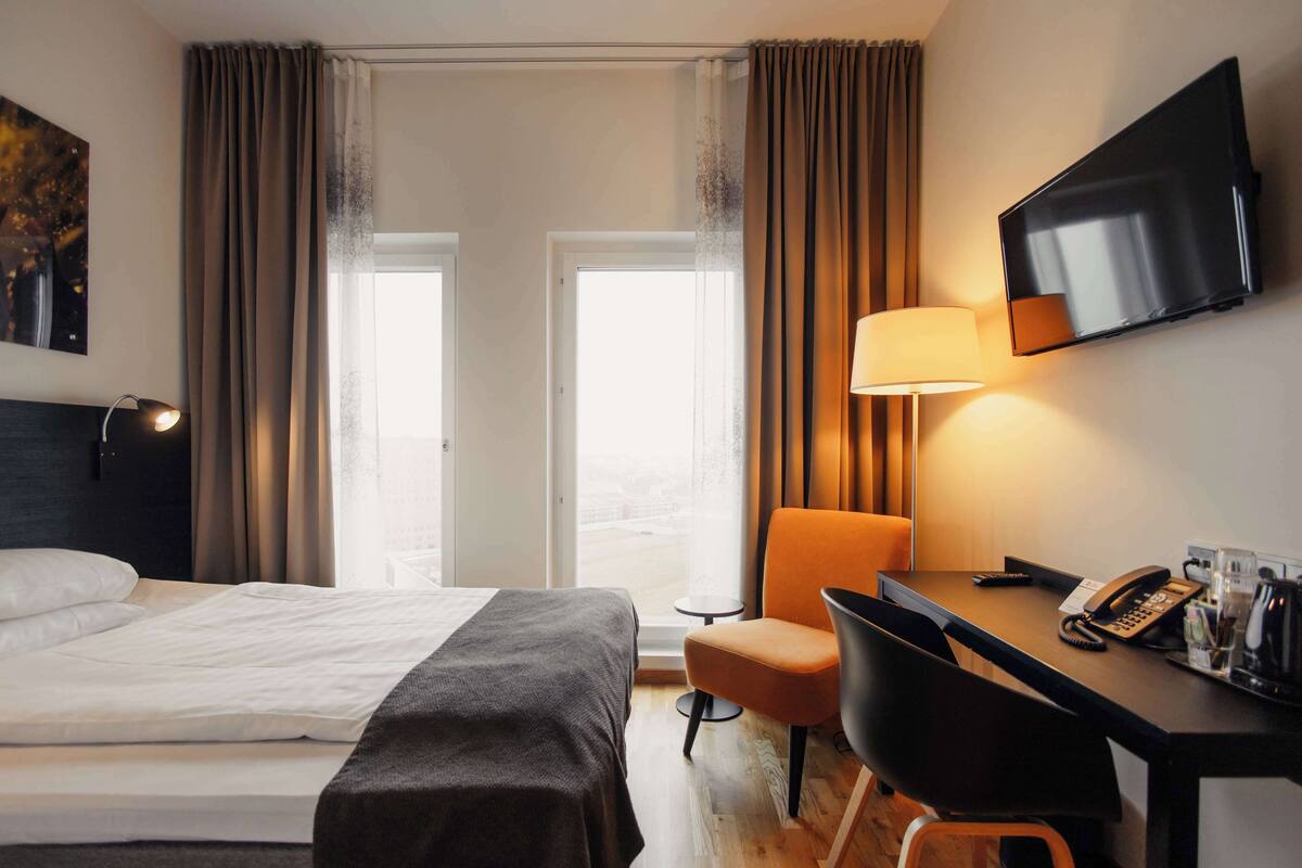 Une chambre d’hôtel avec un lit, un bureau, une chaise, une télévision et une fenêtre avec des rideaux.