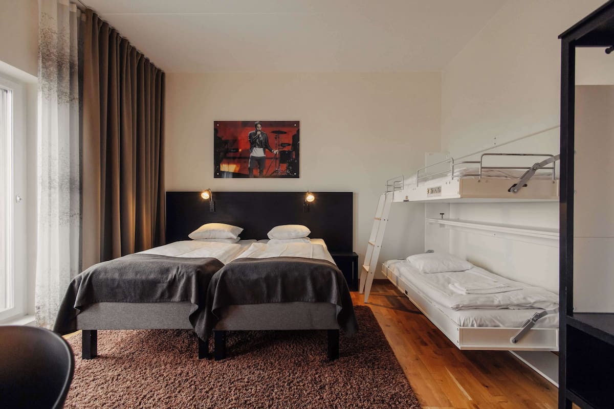 Une chambre avec un lit double, un lit superposé, une grande fenêtre avec des rideaux, un tableau au mur et un sol recouvert de moquette.