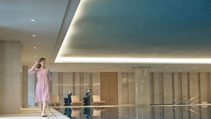 Un área de piscina interior moderna con piso de vidrio transparente, sillas de descanso y techo alto con iluminación empotrada.