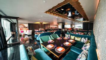 A modern restaurant with teal seating, a central dining table set with orange placemats and purple glassware, and a unique ceiling design.