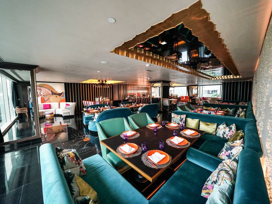 A modern restaurant with teal seating, a central dining table set with orange placemats and purple glassware, and a unique ceiling design.