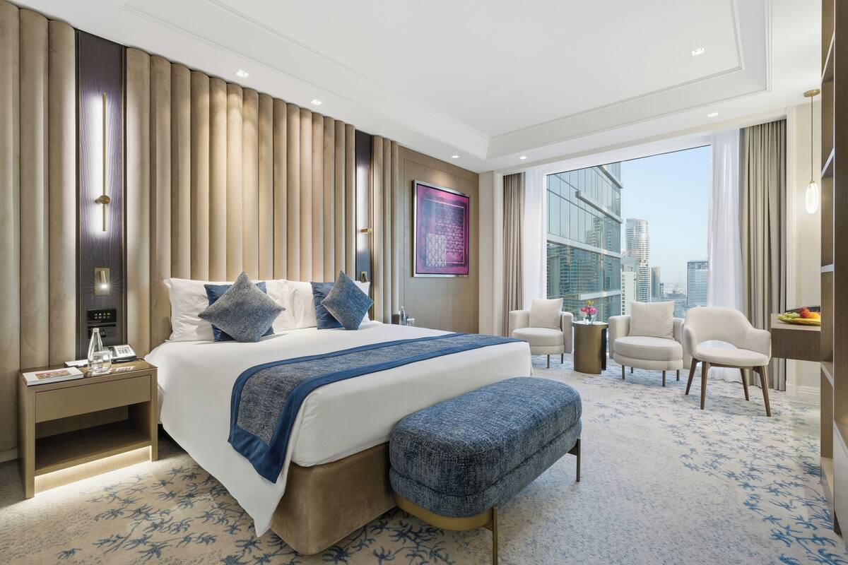 A hotel room with a large bed, a desk, a chair, a small table, and a view of the city.
