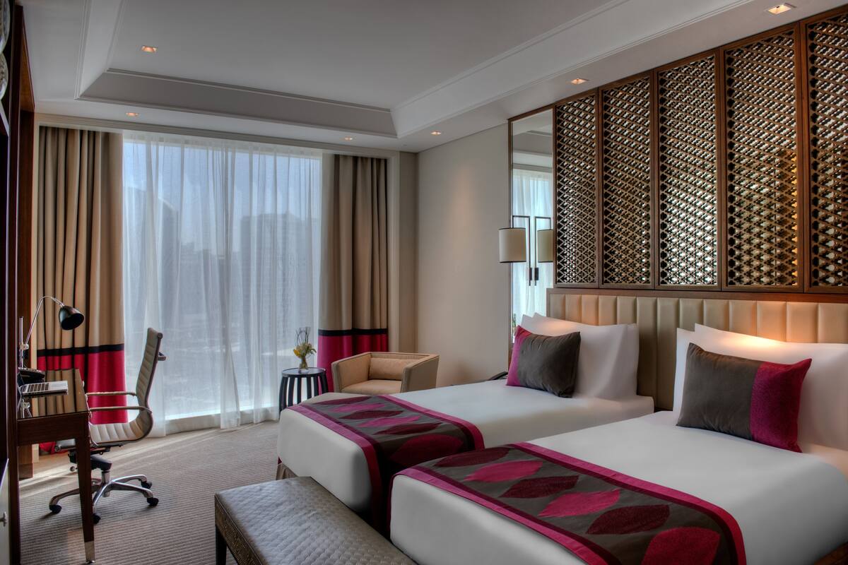 A hotel room with two beds, a desk, a chair, and a large window with curtains.