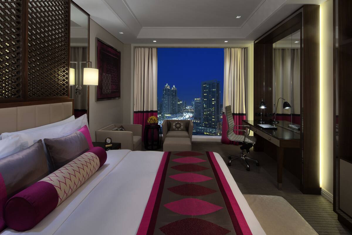 A hotel room with a large bed, a desk, a chair, a sofa, a nightstand, and a view of the city skyline at night.