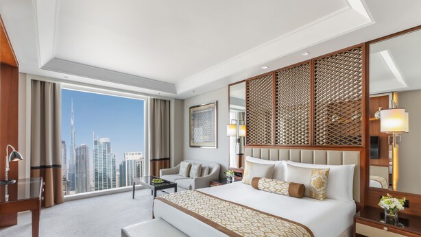 A hotel room with a large bed, a desk, a chair, a bench, a nightstand, and a large window with a city view. - Taj Dubai (Dubai)
