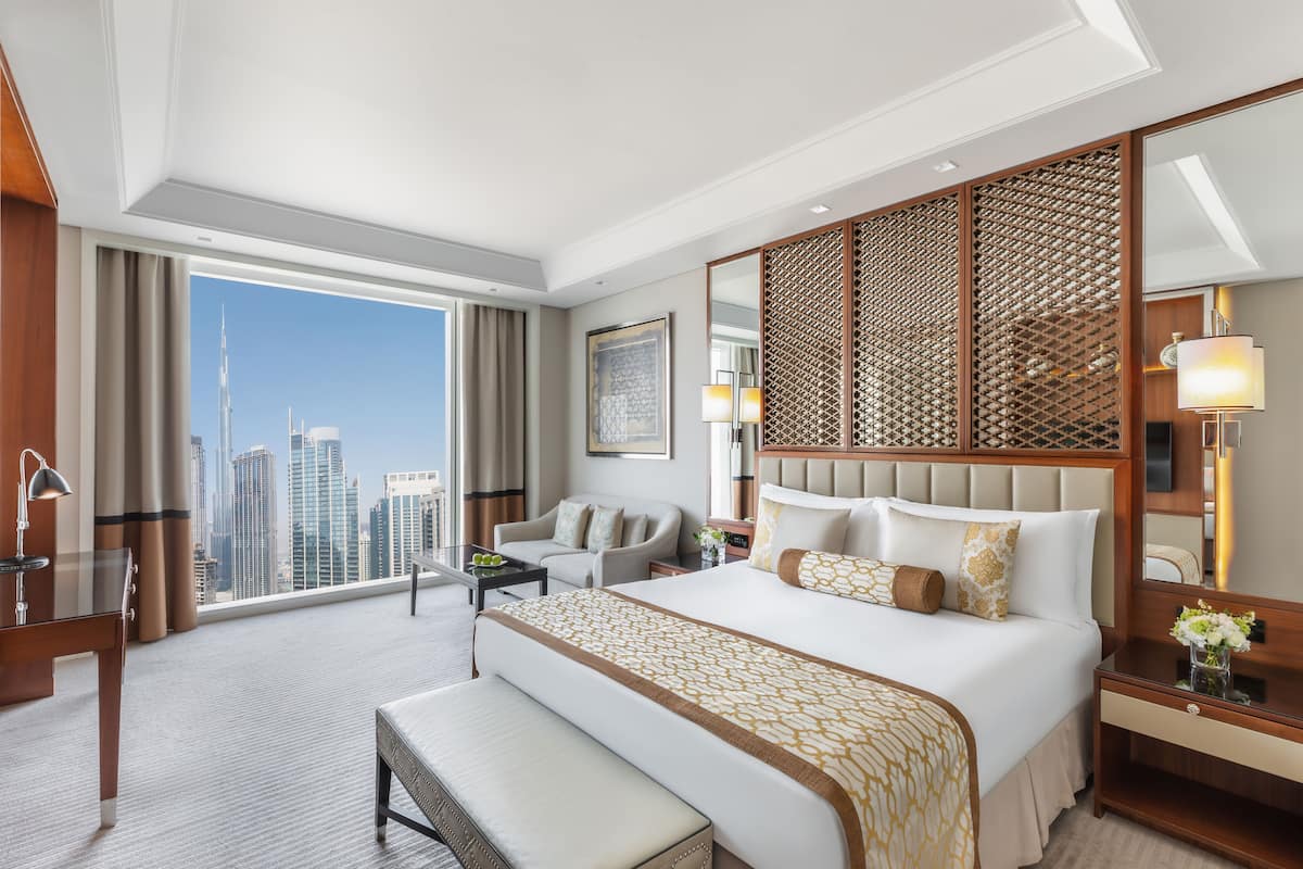 A hotel room with a large bed, a desk, a chair, a bench, a nightstand, and a large window with a city view.