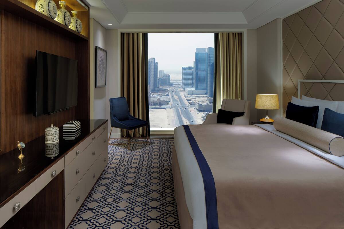 A hotel room with a large bed, a TV on the wall, a desk with a chair, and a view of a cityscape through the window.