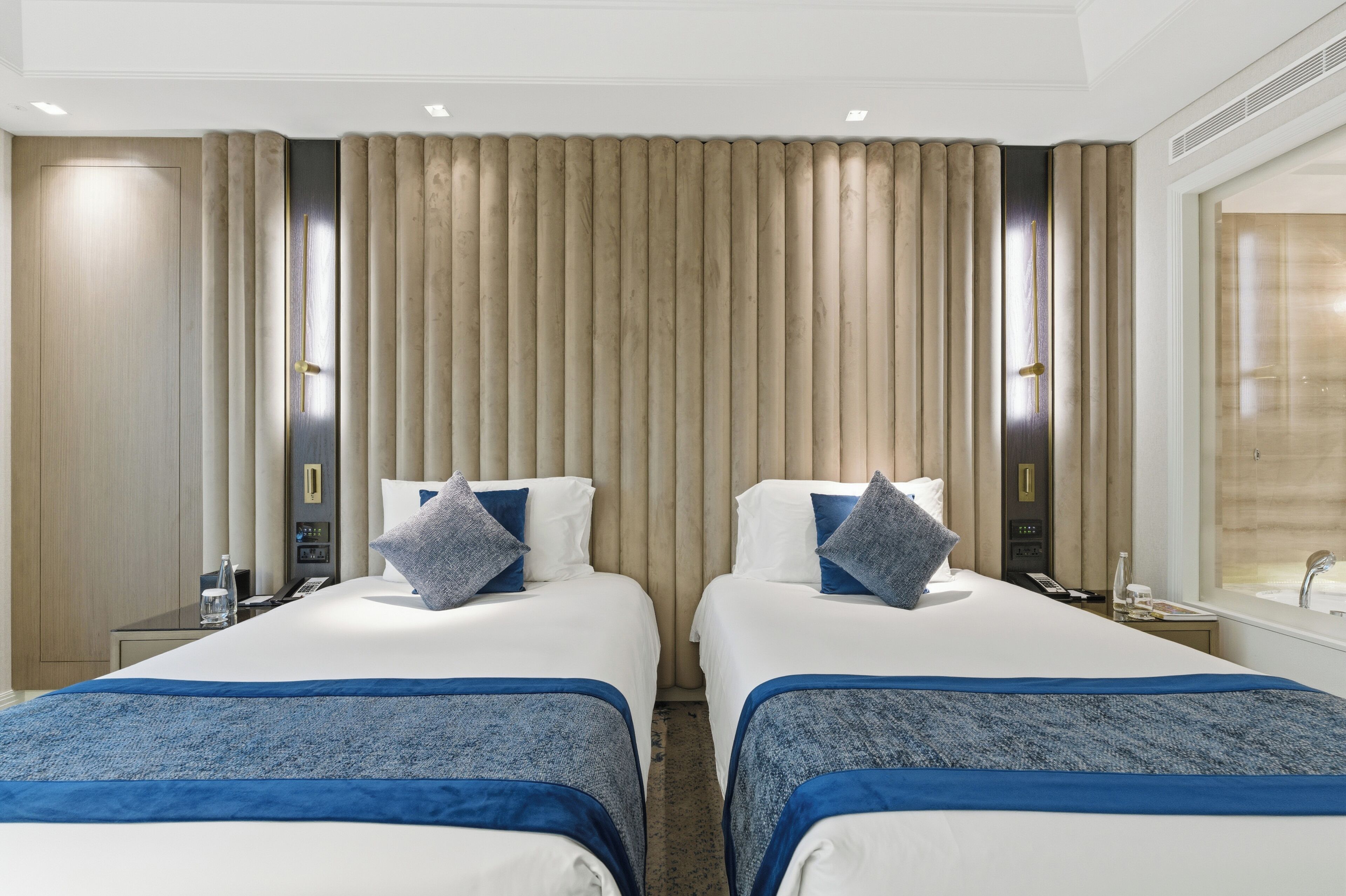 A hotel room with two beds, a minibar, a desk with a phone, and a bathroom with a sink and mirror.