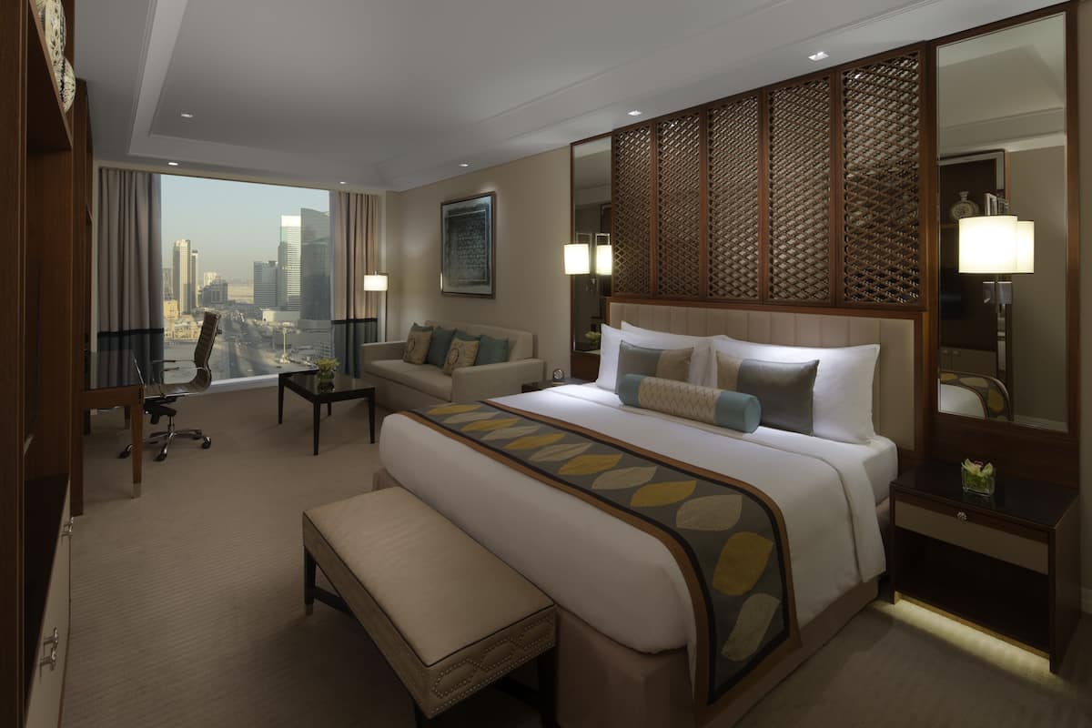 A hotel room with a large bed, a seating area with a sofa and armchairs, a desk, and a large window offering a city view.