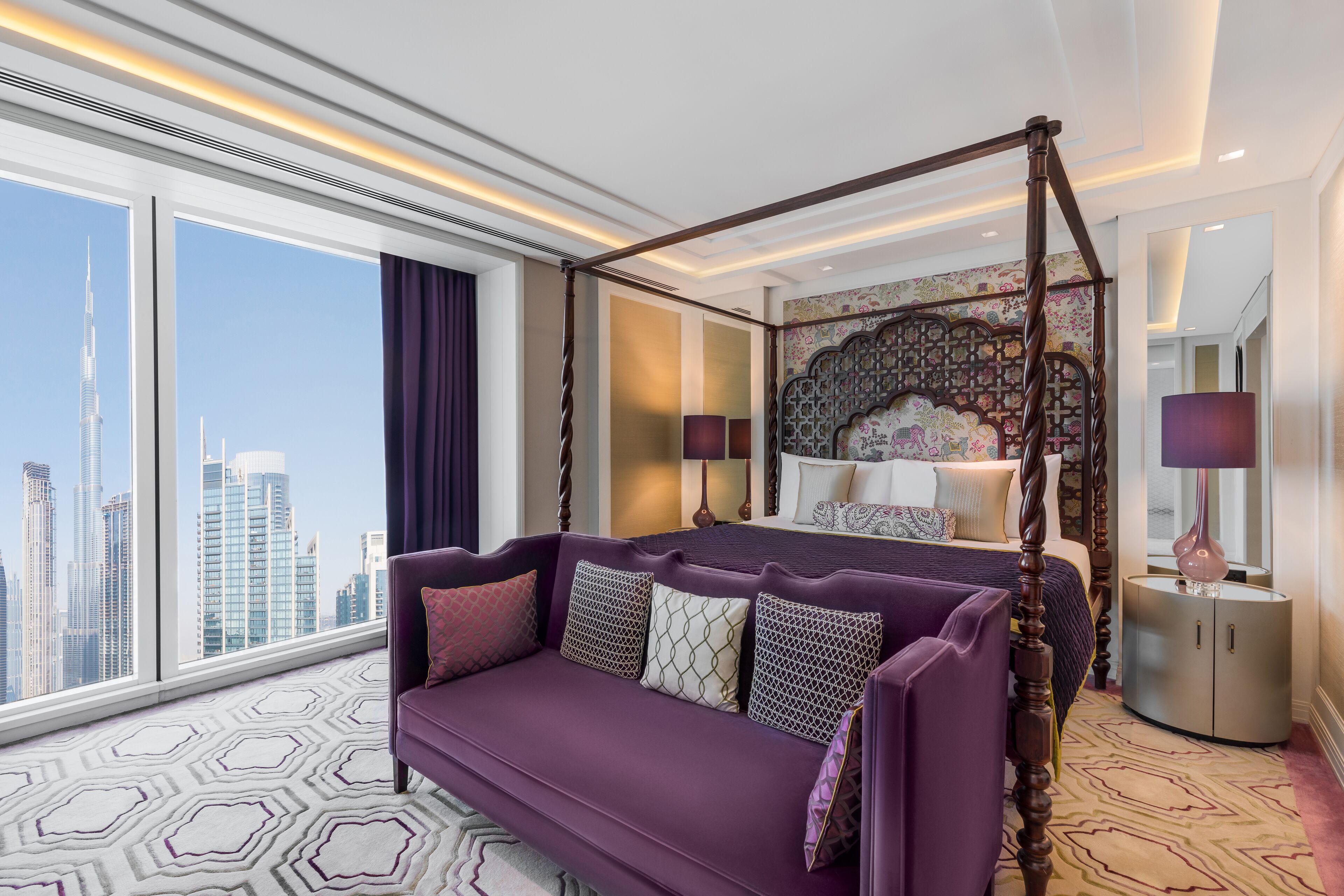 A modern hotel room with a large bed, a purple sofa, and a city view.
