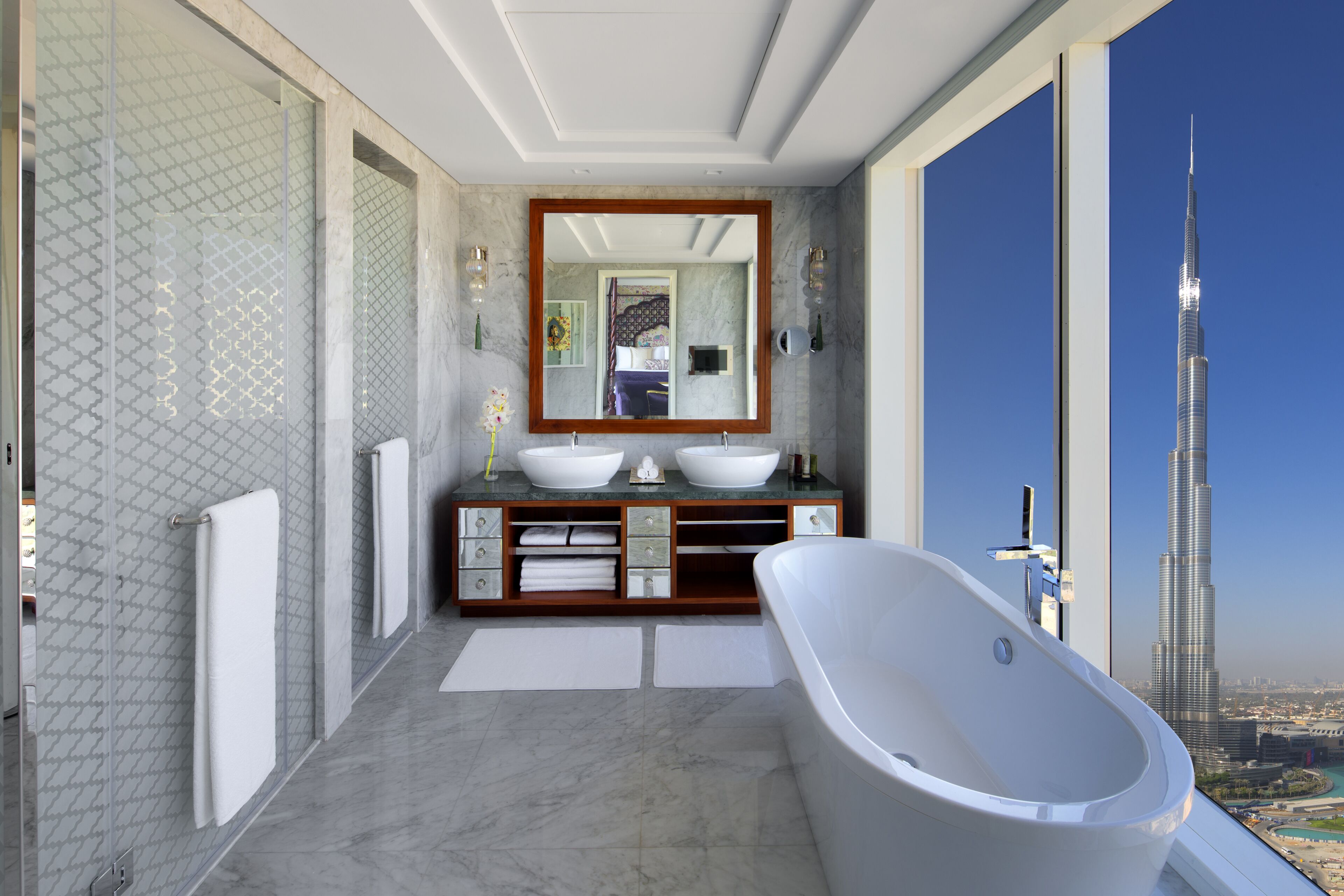 A modern bathroom with a large bathtub, dual sinks, and a large mirror. A view of the Burj Khalifa is visible through the window.