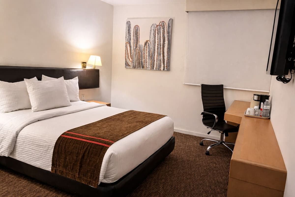 A hotel room with a bed, a desk, a chair, a lamp, and a wall art piece.