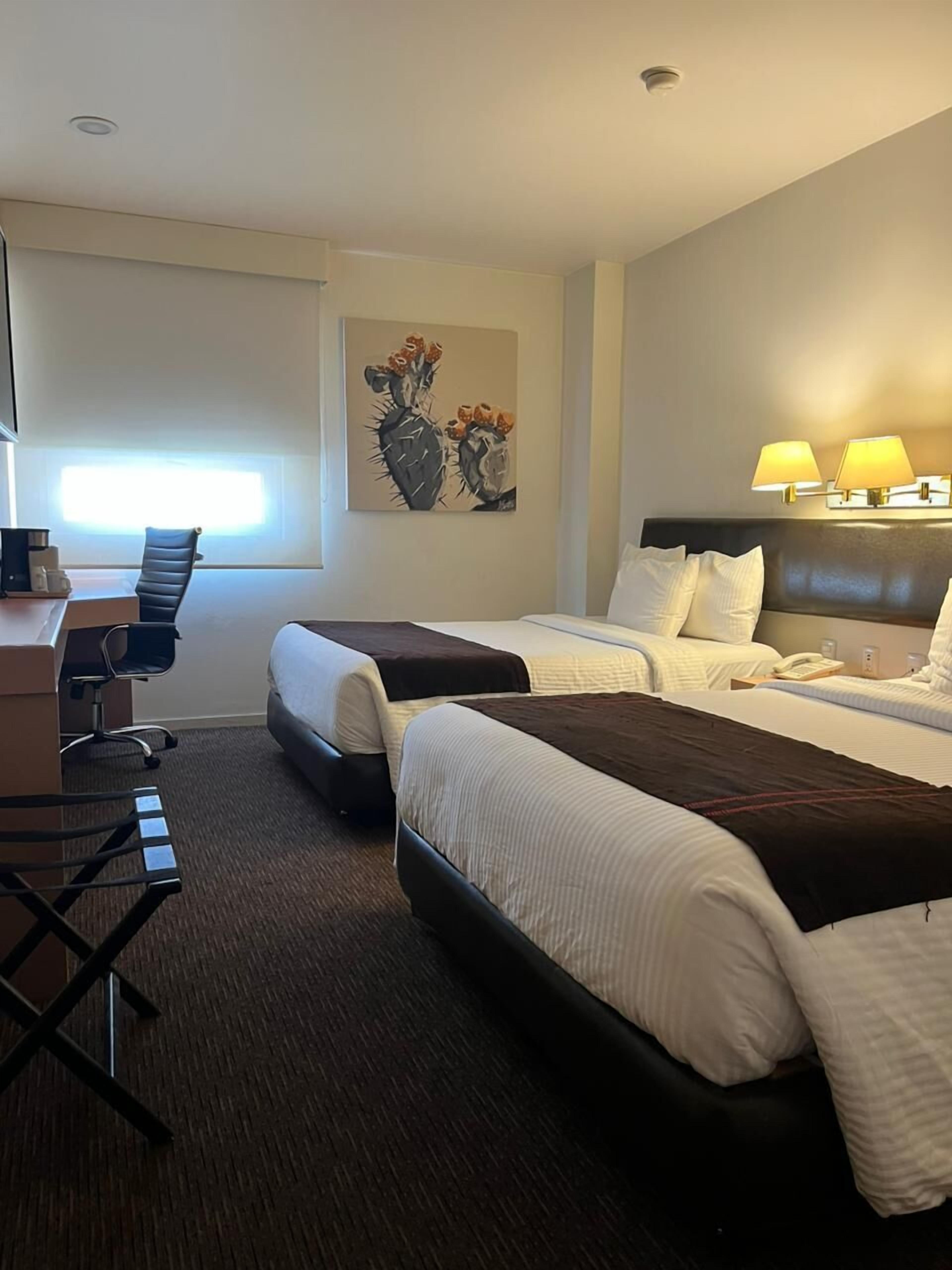 Habitación de hotel con dos camas, un escritorio, una silla, una lámpara y un cuadro en la pared.