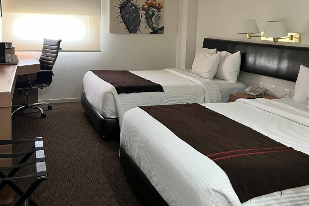 A hotel room with two beds, a desk, a chair, a painting, and a window with blinds.