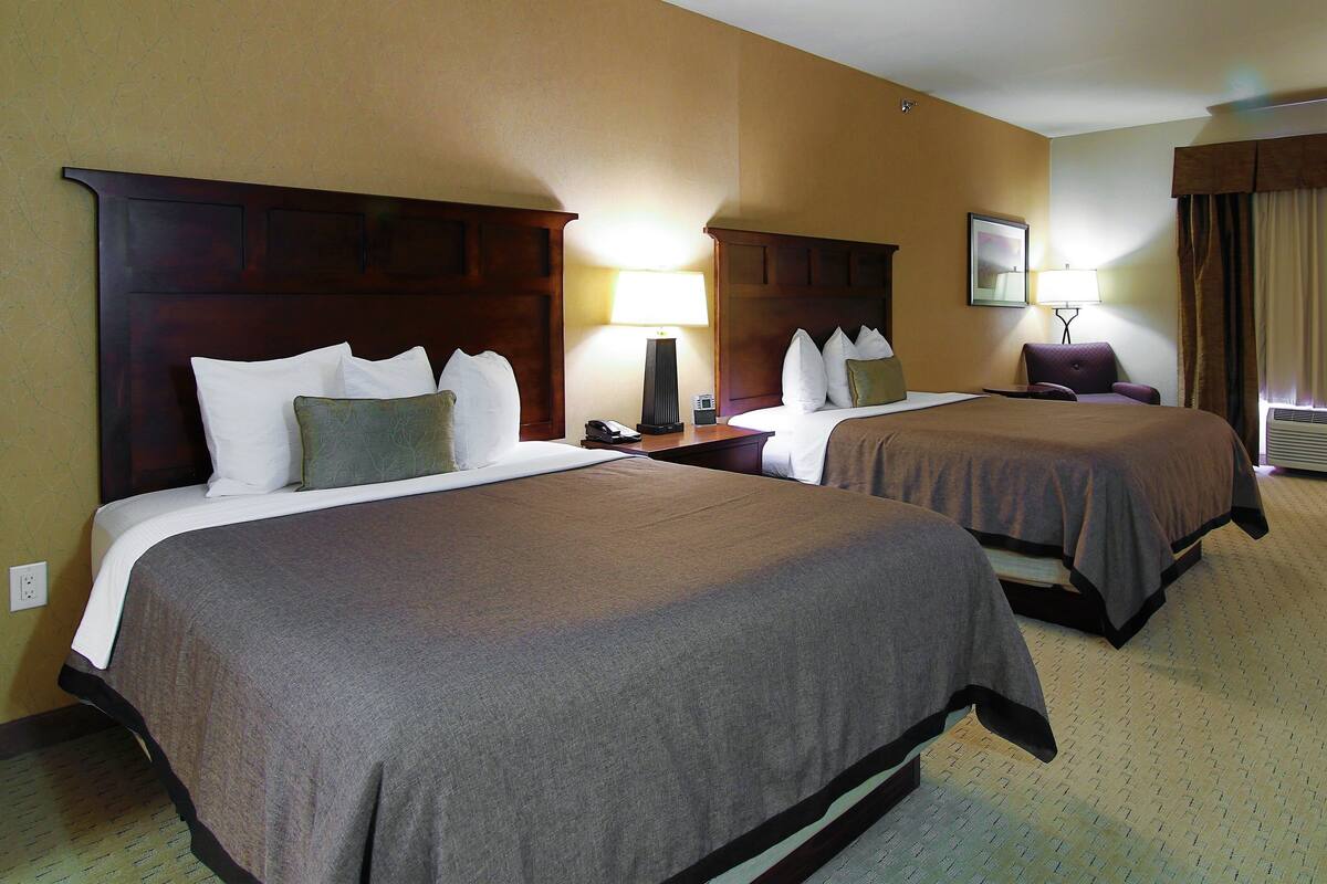 A hotel room with two beds, a nightstand with a lamp, a chair, and a window with curtains.