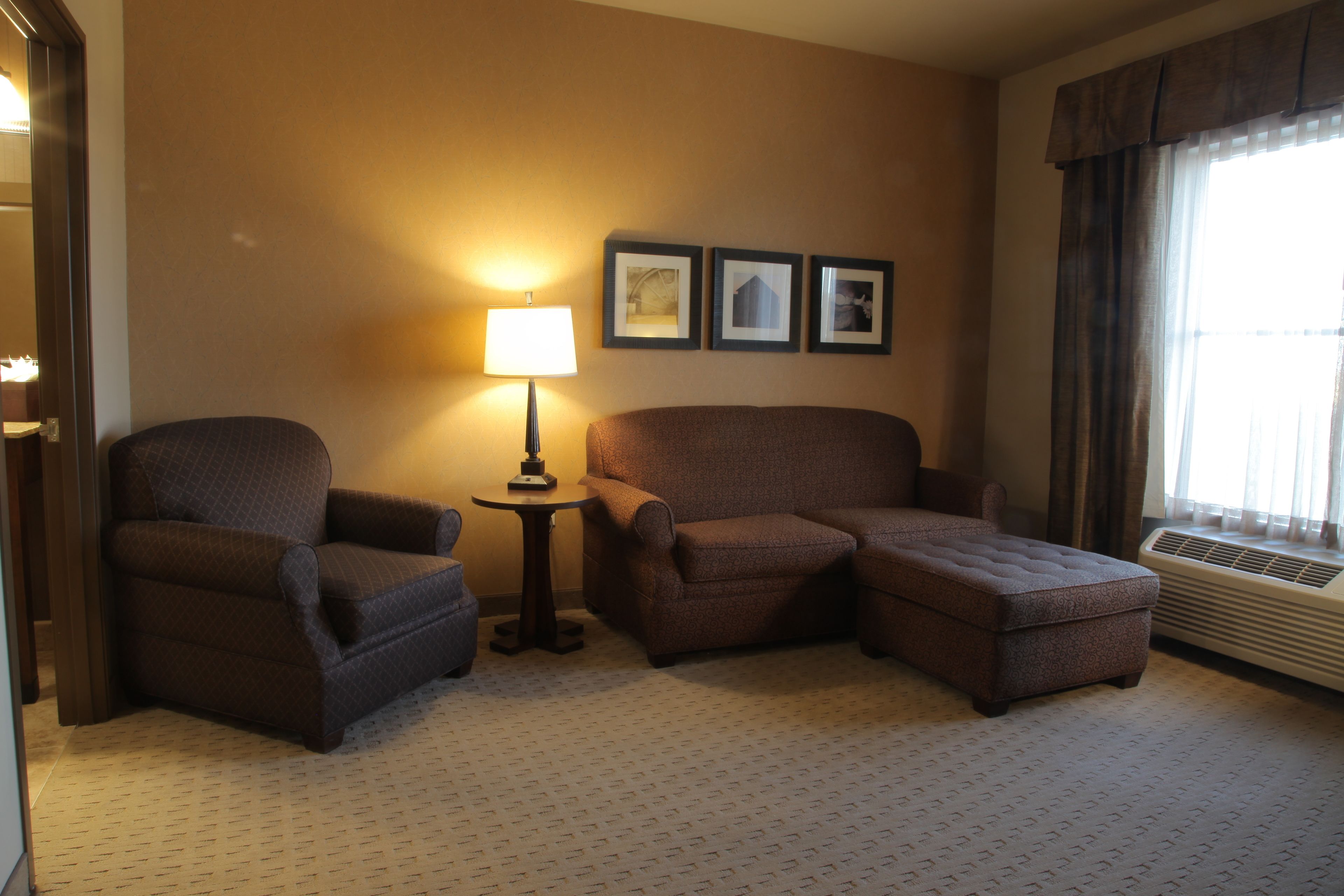 A hotel room with a large brown sofa, a smaller armchair, a side table with a lamp, and three framed pictures on the wall.