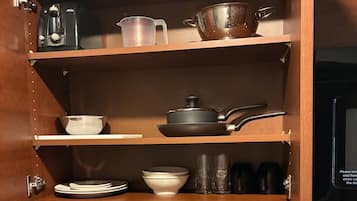 A kitchen cabinet with shelves holding various kitchenware including a coffee maker, measuring cups, a pot, plates, bowls, and cups.