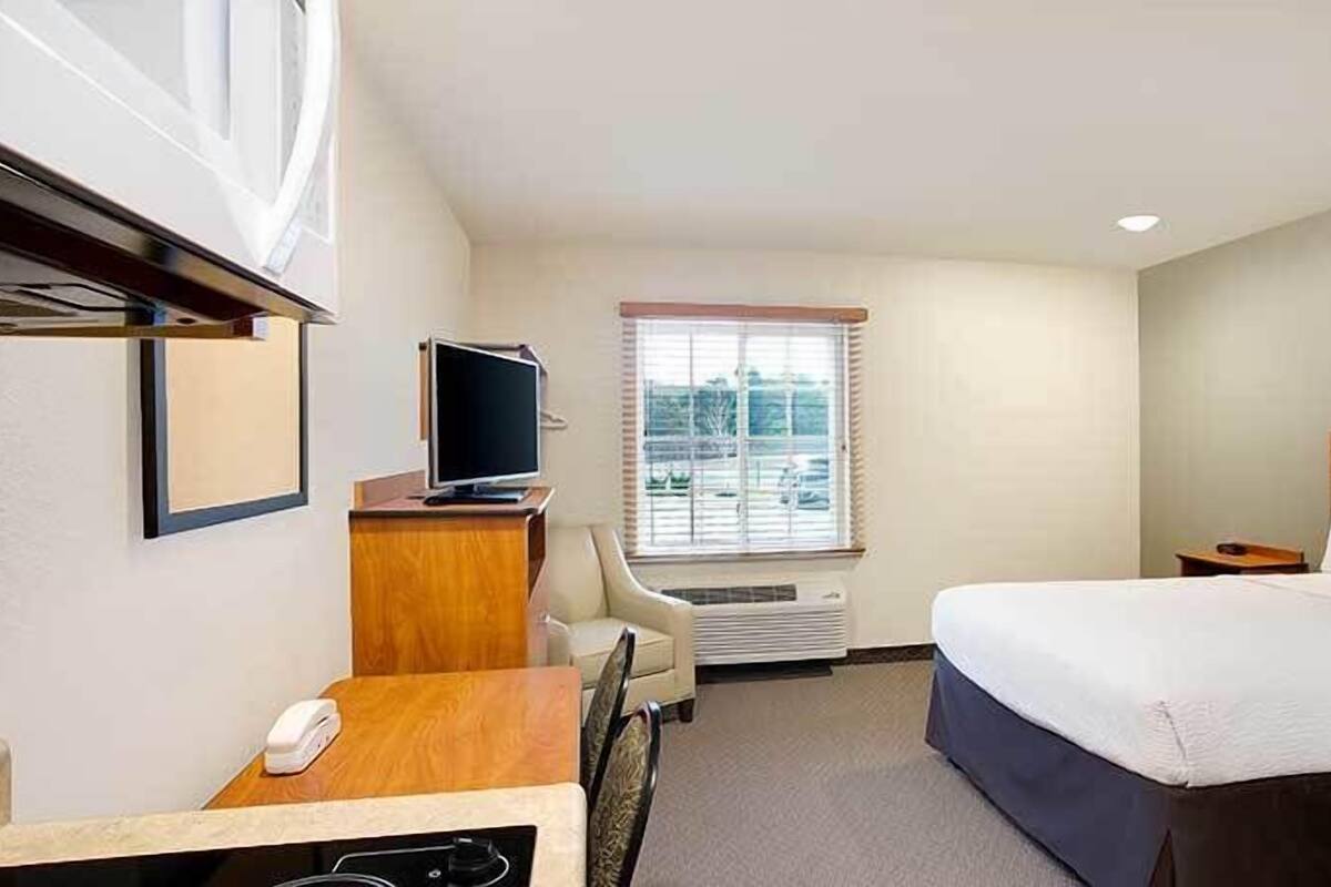 Ein Hotelzimmer mit Bett, Schreibtisch mit Computer, Stuhl, Fernseher und Fenster mit Jalousien.