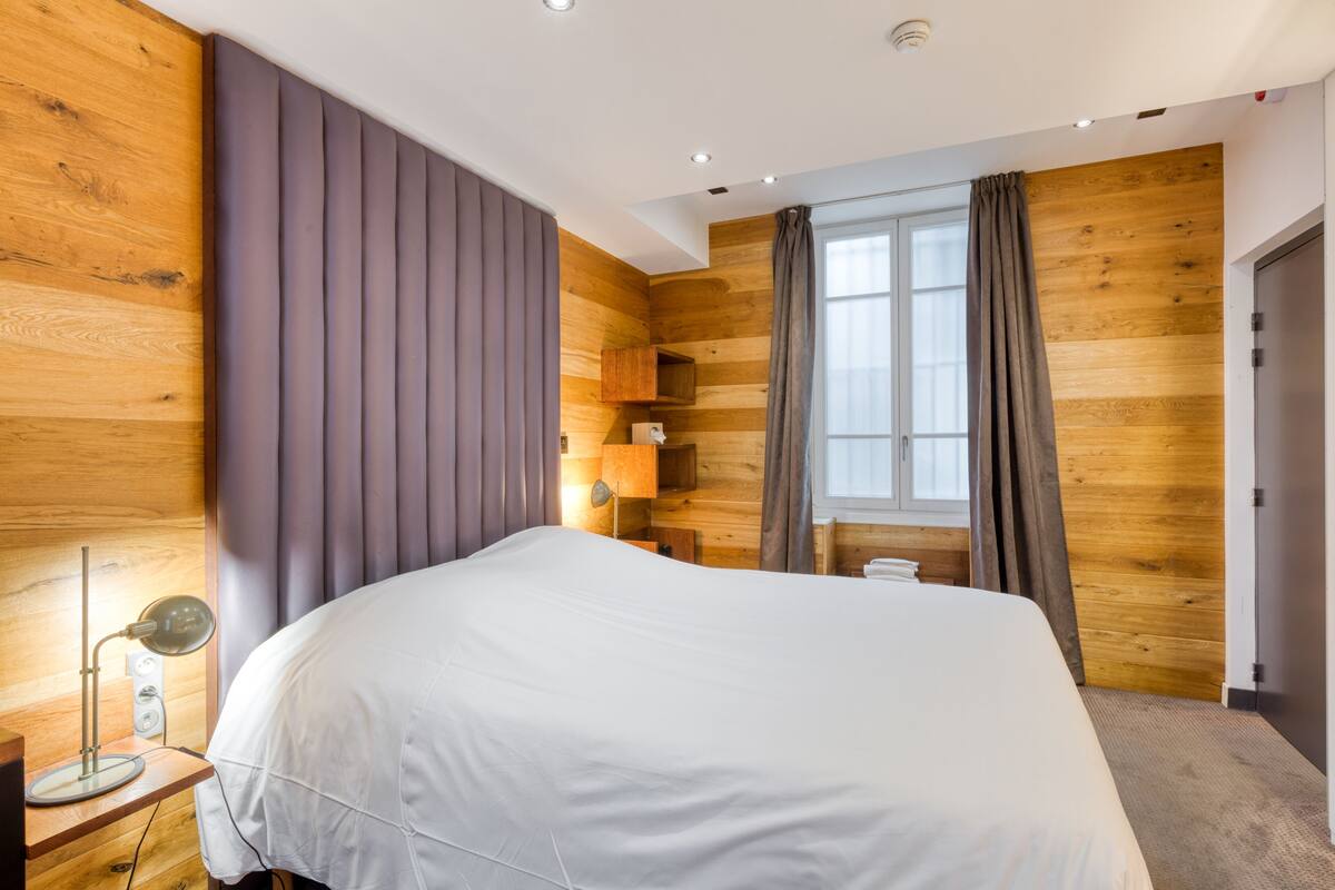 Une chambre d’hôtel moderne avec un grand lit, des murs recouverts de panneaux de bois, une fenêtre avec des rideaux et une lampe de chevet.