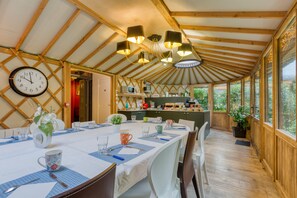 Un espace repas avec une longue table dressée pour le repas, des poutres en bois au plafond et une horloge au mur.