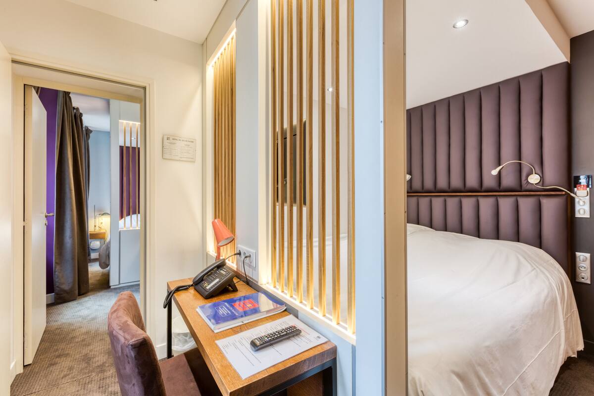 Ein Hotelzimmer mit Bett, Schreibtisch, Telefon, Fernbedienung, Stuhl und einem Fenster mit Jalousien.
