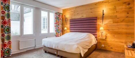 A bedroom with a bed, wooden wall paneling, a window with curtains, and a nightstand with a lamp.