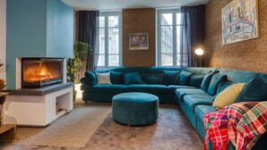Een woonkamer met een blauwgroene bank, een open haard en een groot raam met gordijnen.