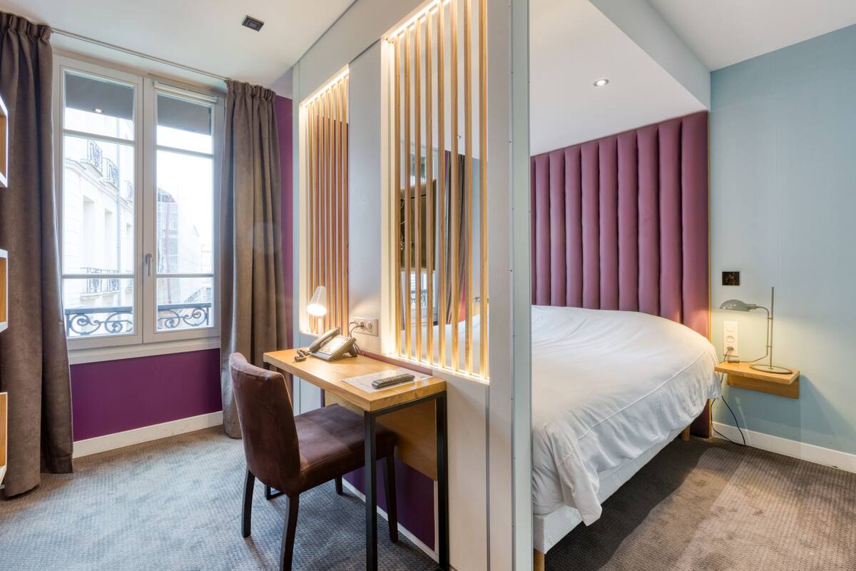 Une chambre d’hôtel comprenant un lit, un bureau avec une chaise, un téléphone et une fenêtre avec des rideaux.