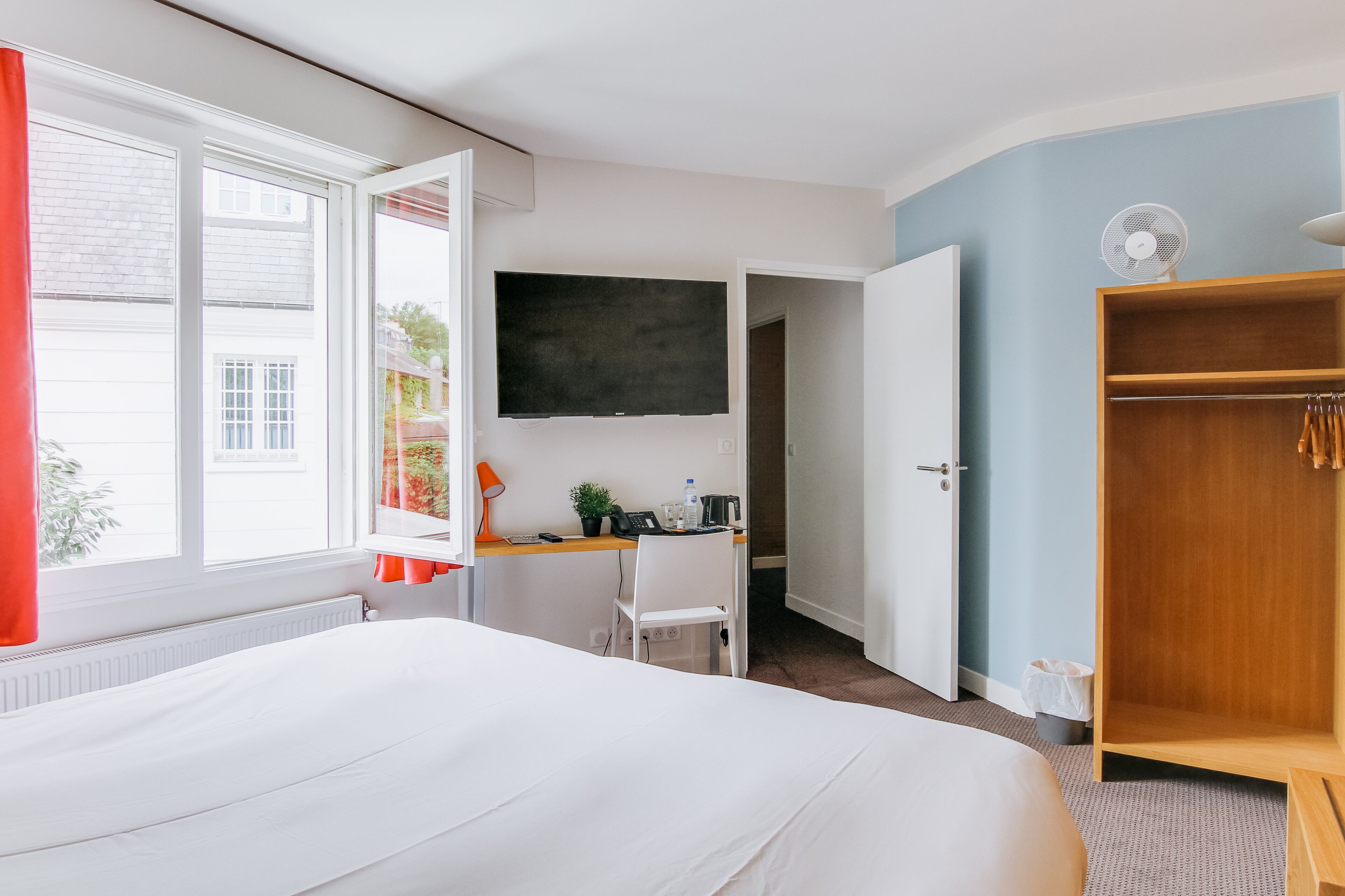 Habitación de hotel con cama, escritorio con silla, televisor, armario y ventana con cortinas.