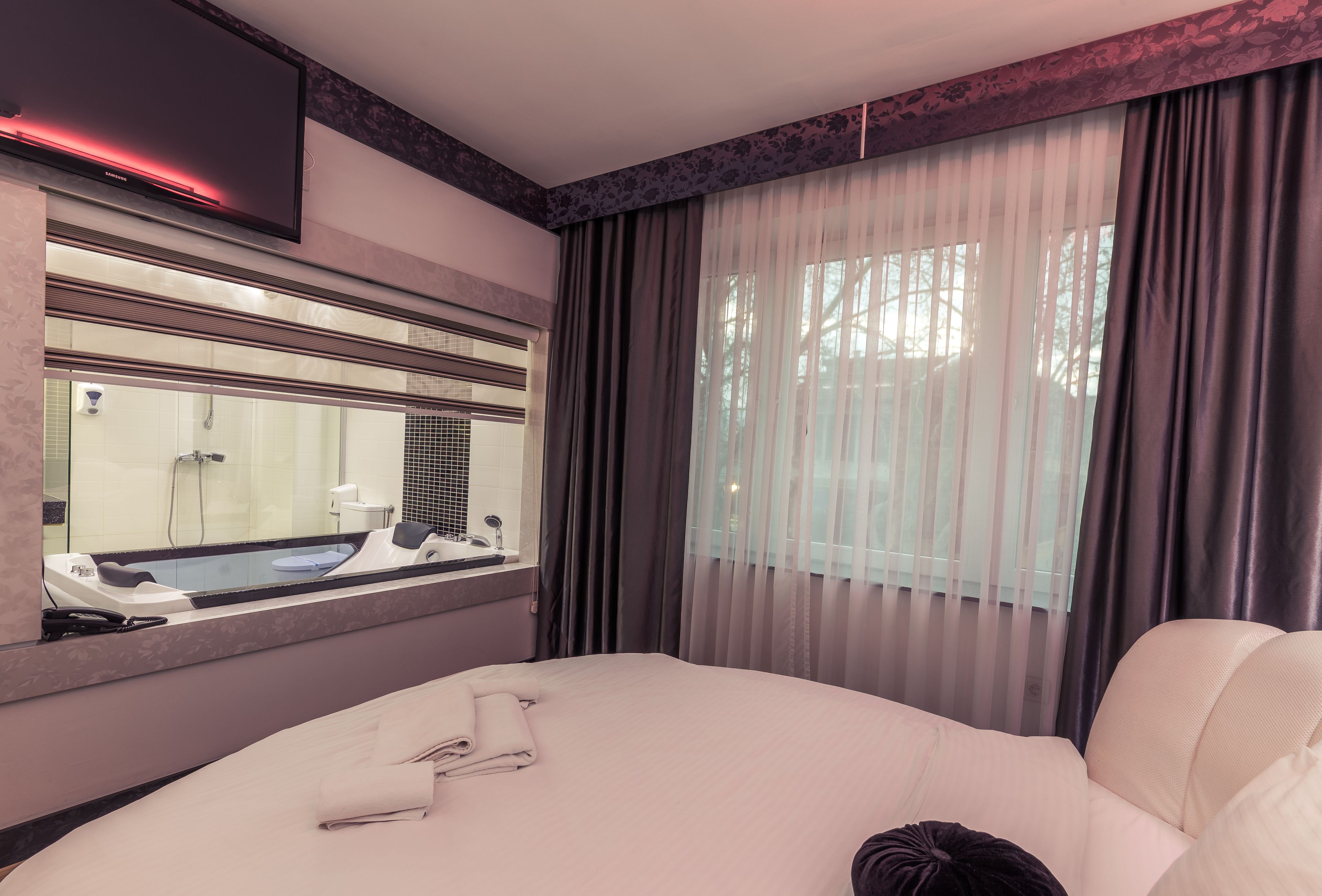 A hotel room with a large bed, a bathroom with a bathtub, a mirror, and a window with curtains.