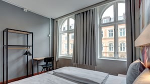 A hotel room with two beds, a desk, a chair, and large windows with curtains.