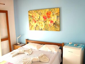 A hotel room with a bed, a nightstand, a painting of flowers, and a minibar. - Pousada Casa das Rosas (Búzios)