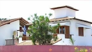 A white building with a brown roof and wooden shutters, surrounded by greenery. - Pousada Casa das Rosas (Búzios)