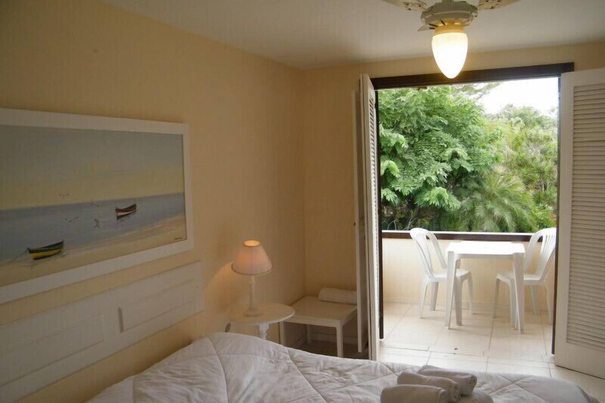 A hotel room with a bed, a painting, a lamp, a small table, and a view of a balcony with chairs.
