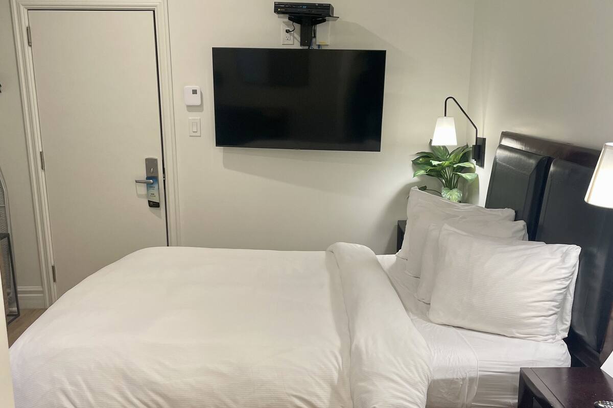 Ein Hotelzimmer mit einem Bett, einem an der Wand montierten Flachbildfernseher, einem Nachttisch mit Lampe und Holzboden.