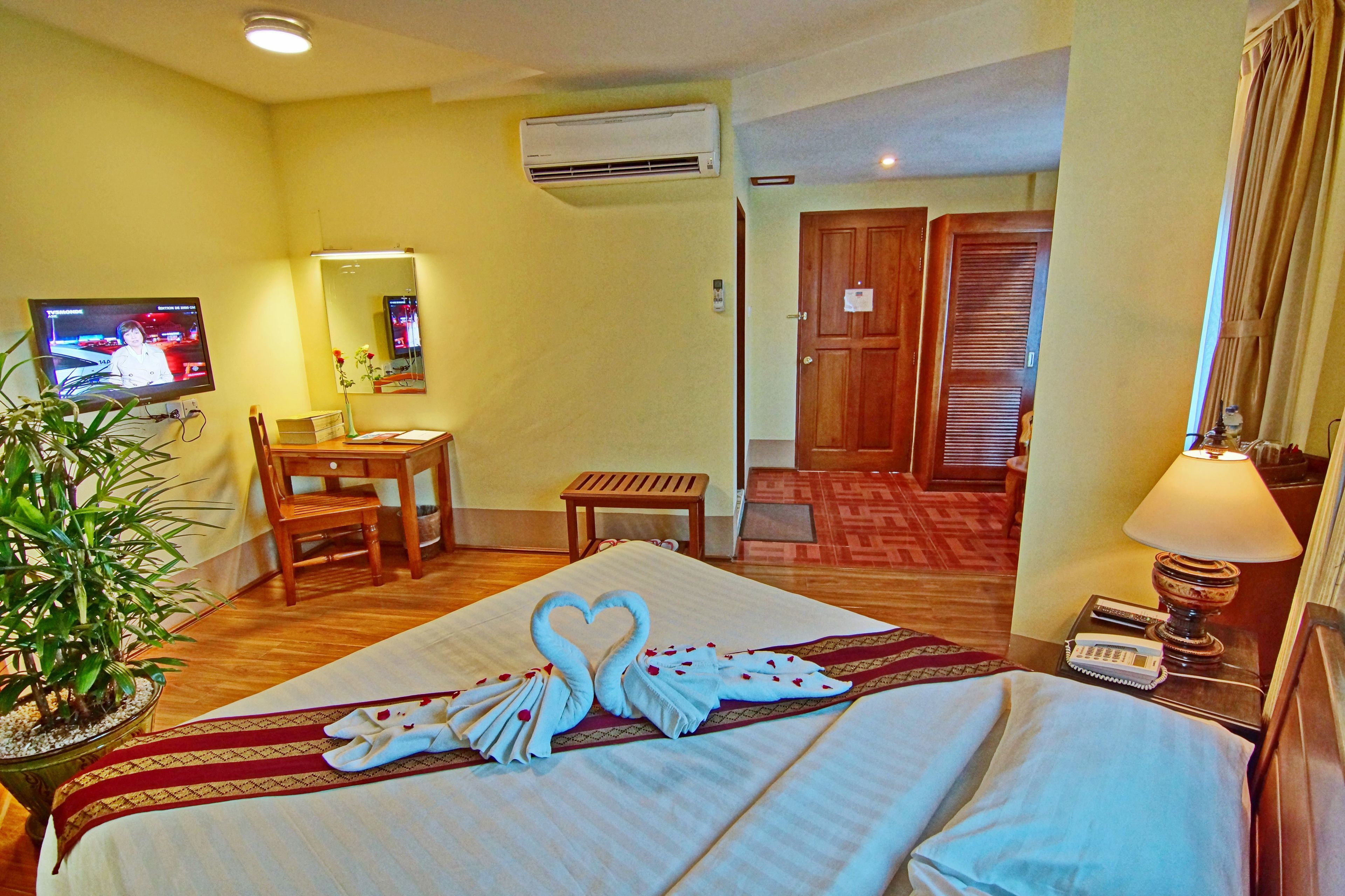 A hotel room with a neatly made bed, a television, a desk with a chair, a small table, and a potted plant.
