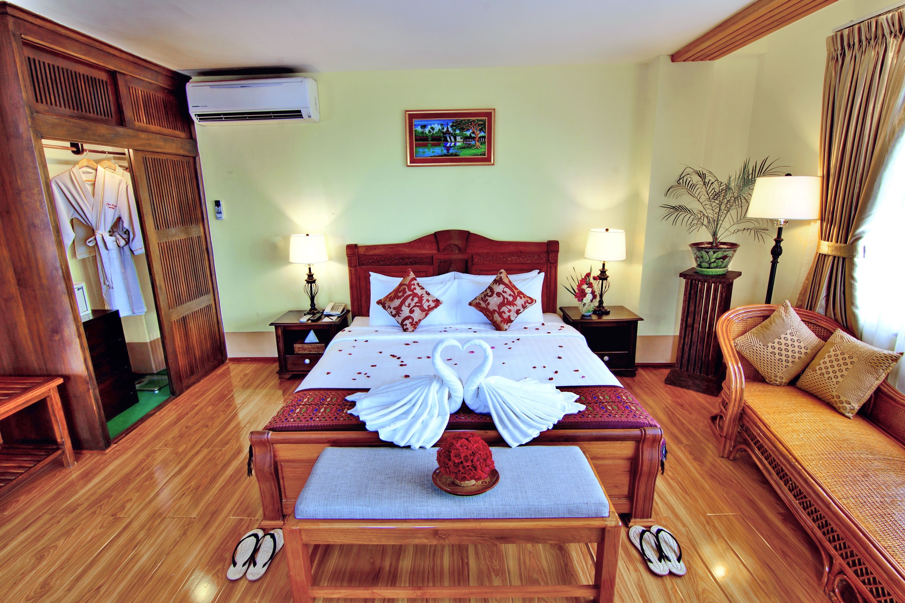 A hotel room with a large bed, wooden furniture, a potted plant, and a painting on the wall.