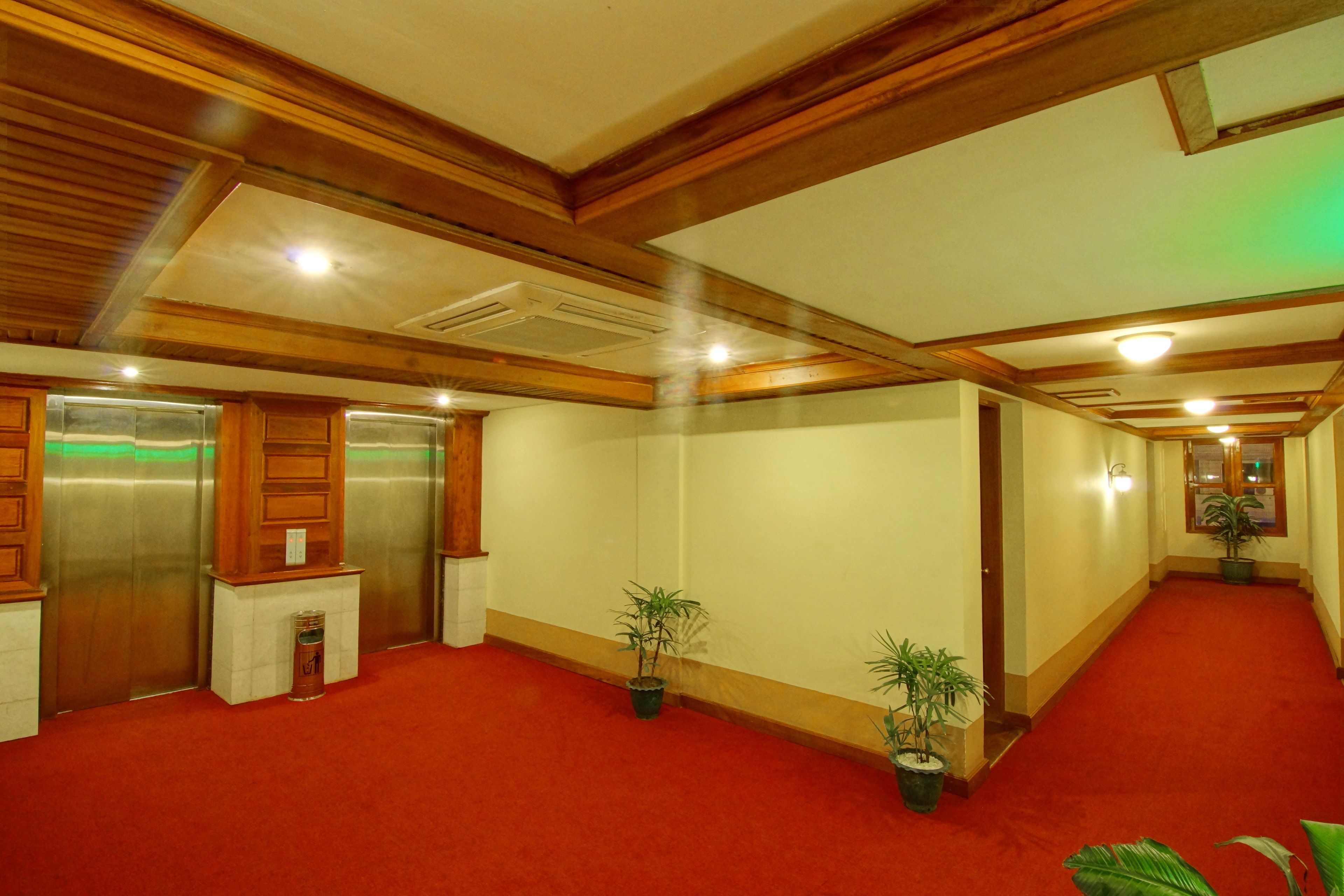 A hotel corridor with red carpet, wooden ceiling beams, and potted plants.
