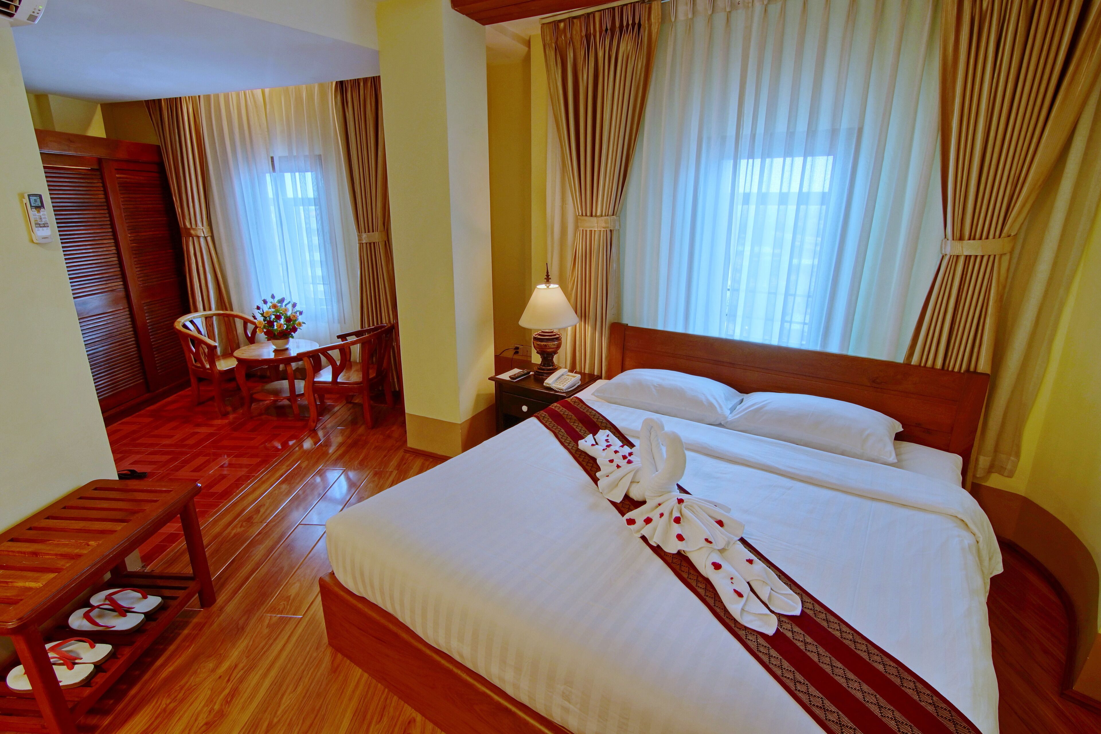 A hotel room with a large bed, a wooden bench with slippers, a small table with a vase, and a chair.